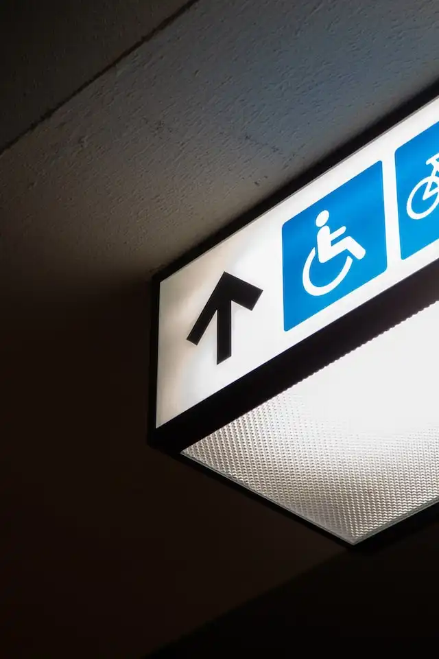 Illuminated accessibility sign with symbols for an elevator for disabled access and bicycle parking, featuring an upward arrow against a dark ceiling.