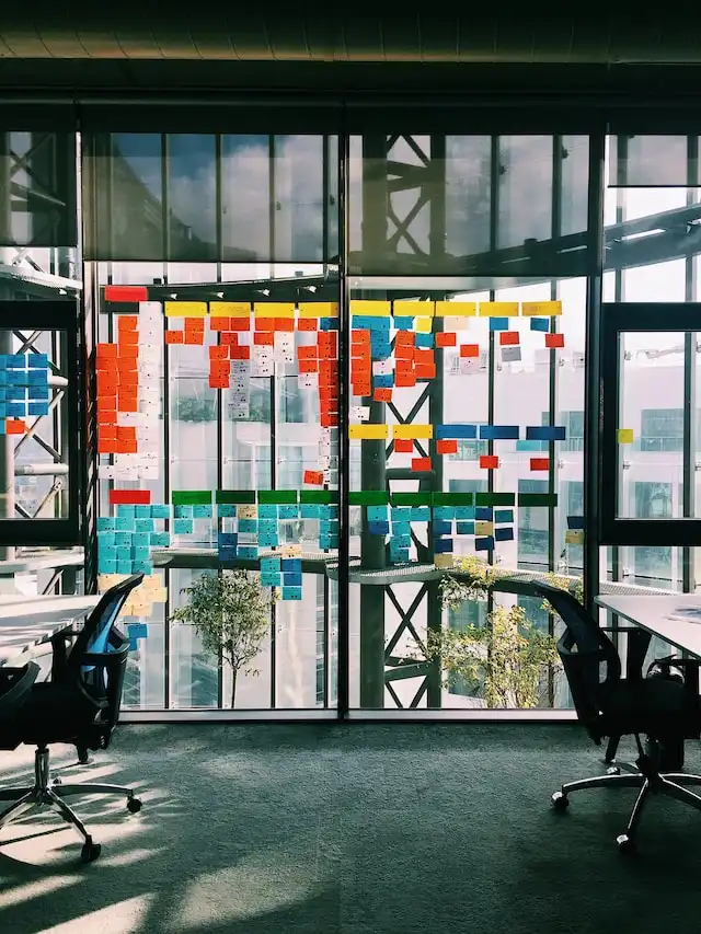 Office space with two black chairs facing a glass wall covered in colorful sticky notes arranged in columns for agile planning.