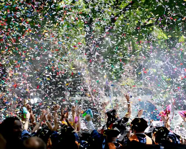 A jubilant crowd of graduates celebrates their commencement. The air is filled with a vibrant shower of confetti, reflecting a spectrum of colors that glisten in the light. Graduation caps are tossed up in joy, contributing to the festive atmosphere.