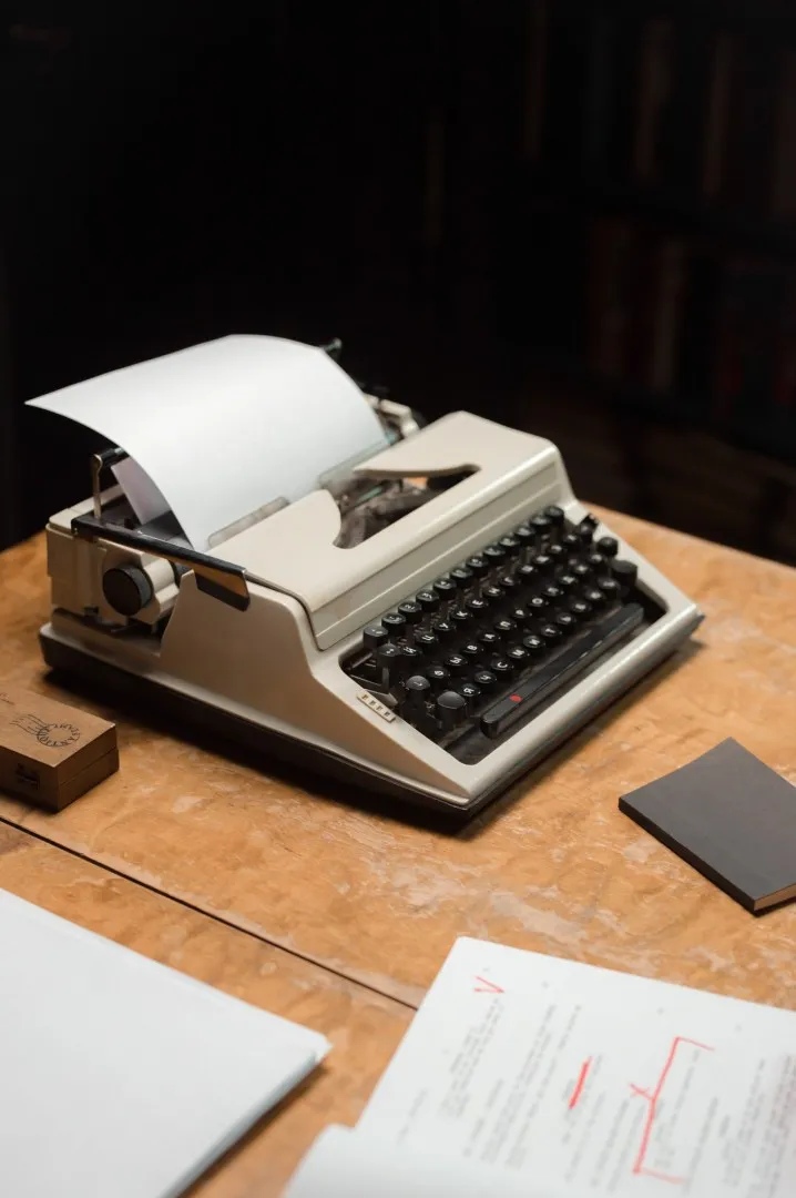 A vintage typewriter with a sheet of paper inserted, ready for typing. The typewriter is placed on a wooden table top with a small black notebook and other materials nearby.