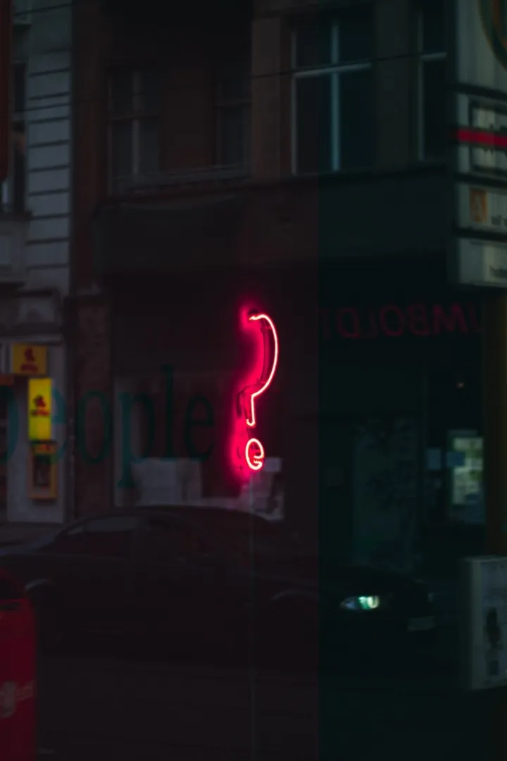A red neon sign in the shape of a question mark is shown as a reflection with an urban city backdrop.