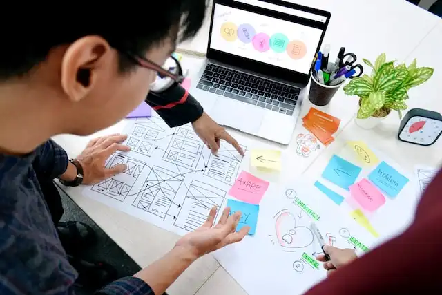Multiple people are engaged in a collaborative design session, with one pointing to a wireframe layout on a white paper and the other drawing a diagram with circular elements and text annotations. The scene is a typical setup for design brainstorming, with tools such as colored sticky notes, pens, and other office supplies scattered around, contributing to a creative and organized workspace.